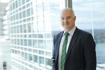 a man in a dark suit stands by a huge window with a glass-panelled building behind him