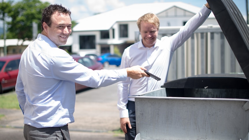 A man prepares to put his smartphone in a rubbish dumpster while another man holds the lid open.