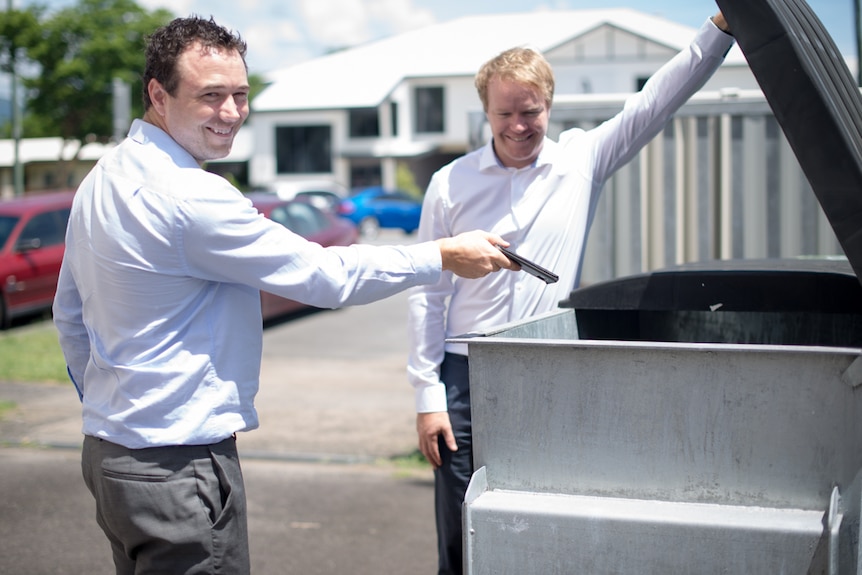 A man prepares to put his smartphone in a rubbish dumpster while another man holds the lid open.