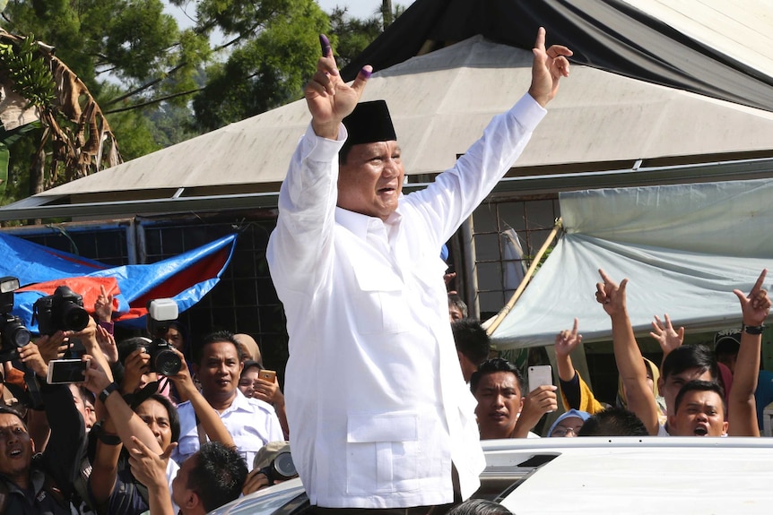 Prabowo Subianto stands out of a car and holds his hands up to the crowd who cheer and take photos.