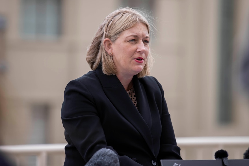 A woman with blonde hair wearing a black jacket speaks to media.