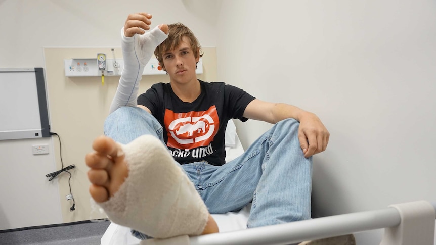 A boy sits with his arm and leg in a cast, his foot is missing his big toe, and the big toe has been attached in place of thumb