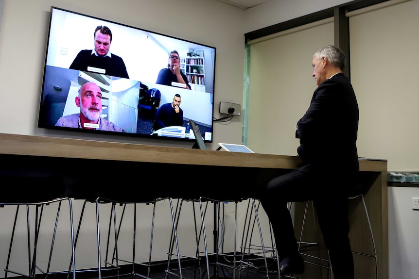 Man wearing suit stands at desk looking at television conference screen divided into four with three men and one woman