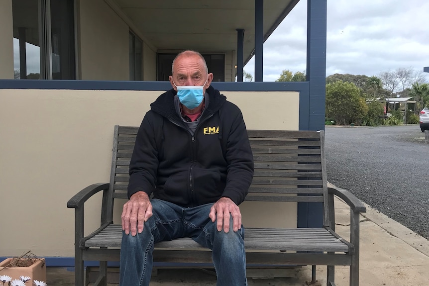 An old man wearing a mask sits on a bench out the front of a motel.