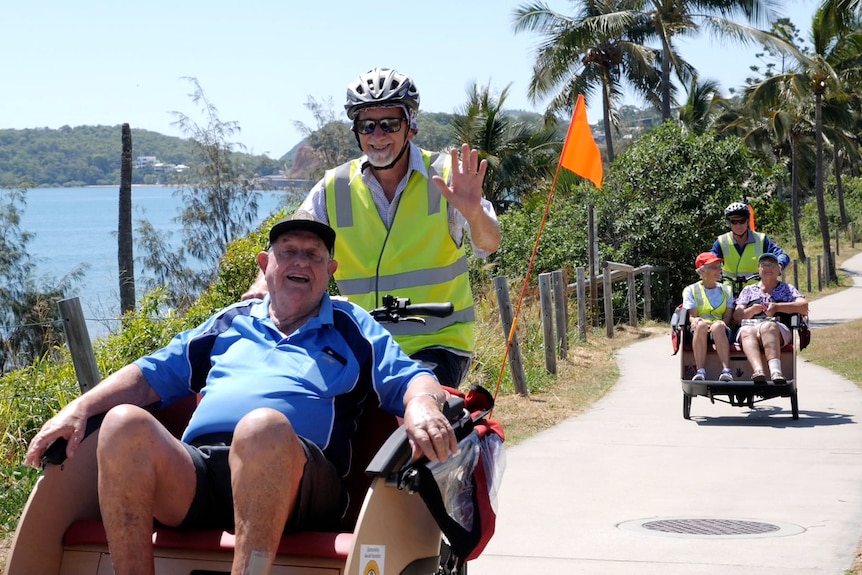 Keith on a trishaw smiling, volunteer waving, Estelle on another trishaw behind.