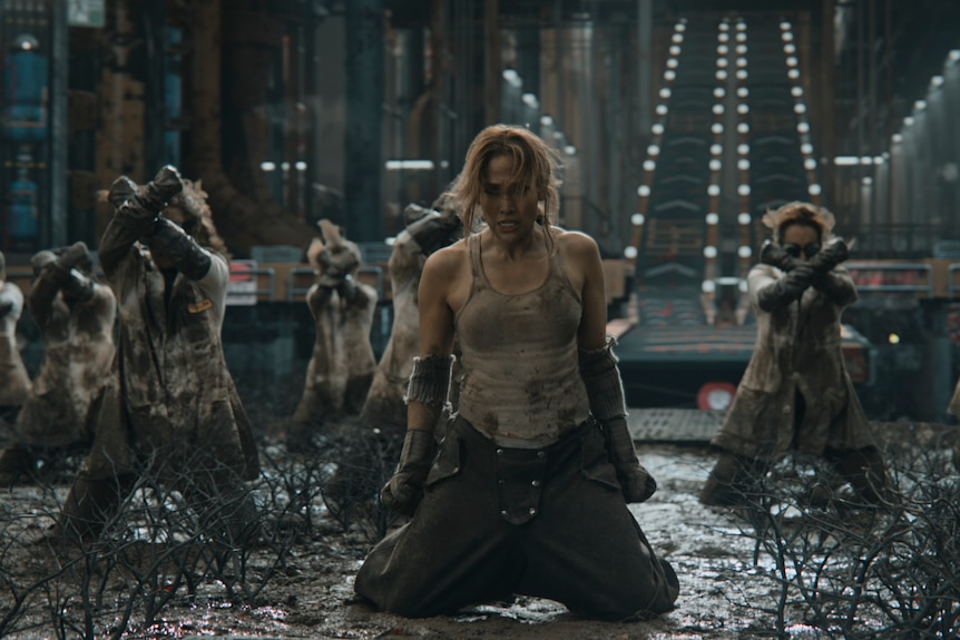 Jennifer Lopez mid dance-sequence on her knees in the mud of a dark dystopian set.