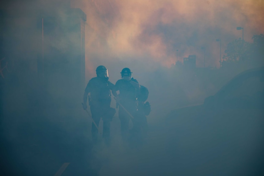 Two police officers are seen engulfed in a cloud of tear gas, the sky above them is an orange haze.