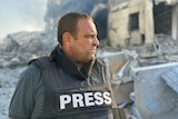 A man stands in front of piles of rubble while wearing a bullet proof vest that says Press on it.