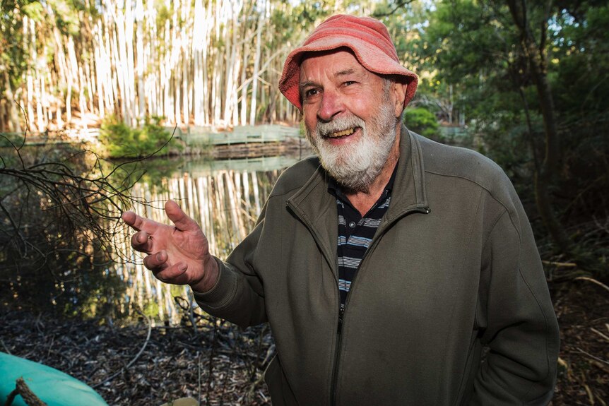 A man in a bucket hat and zip up jersey smiles and gestures with he hands as he stands talking in a nature reserve.