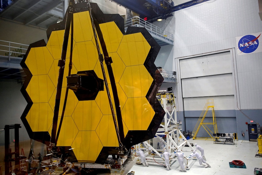 The James Webb Space Telescope Mirror sits in a NASA warehouse comprised of many hexagonal, gold coloured mirrors.