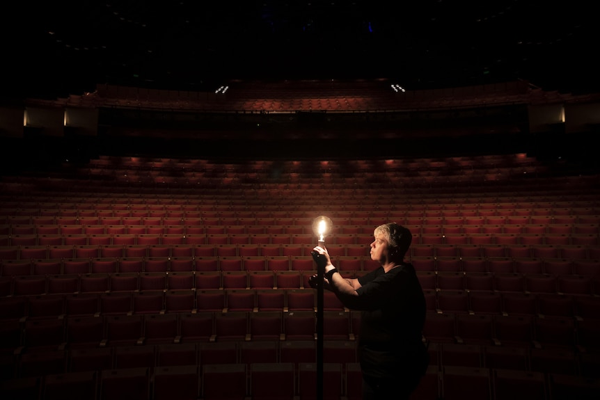 A woman stands in a deserted theatre setting up a lightbulb on a pole.
