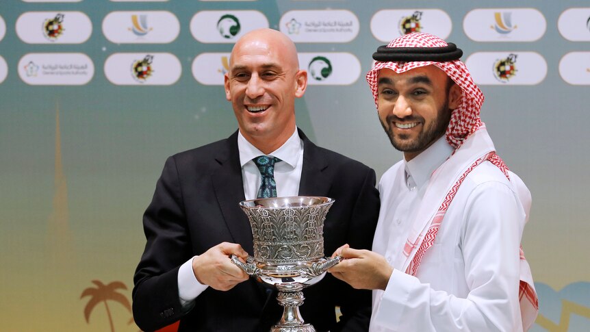 Two men, one wearing a suit, one a white robe, holding a trophy and smiling