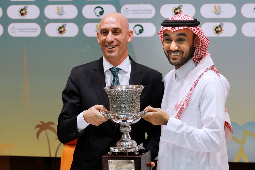 Two men, one wearing a suit, one a white robe, holding a trophy and smiling
