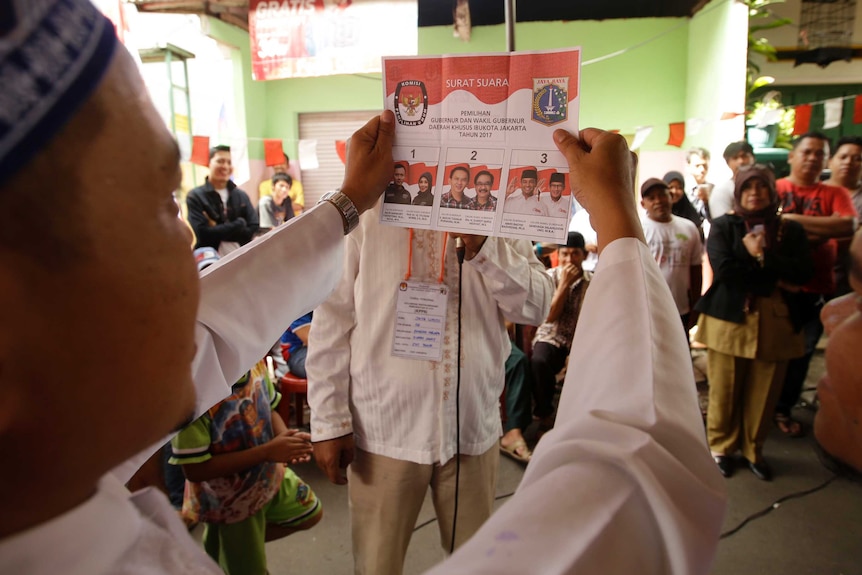 A side profile of a man holding the ballot paper for the election, people in the background look at him.