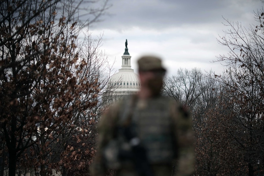 An out-of-focus person wearing a military uniform and a face mask foregrounds the US Capitol Building.