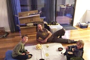 A man and two children eat dinner on a blanket on a floor
