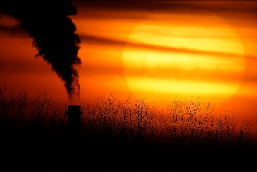 Smoke pours out of a chimney in front of an orange sunset