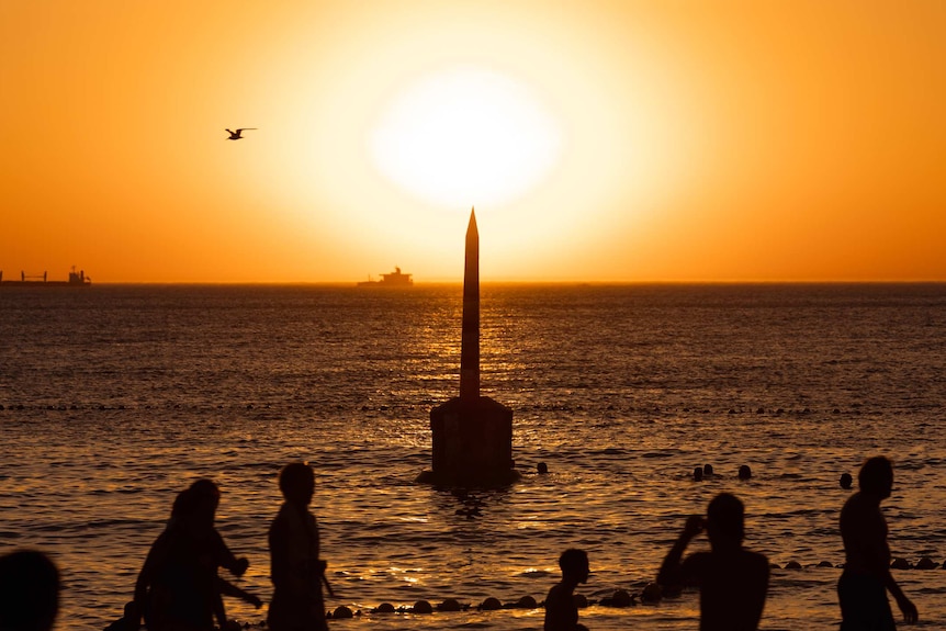 People in silhouette in front of the pylon as the sun sets over at Cottesloe Beach on a hot day.