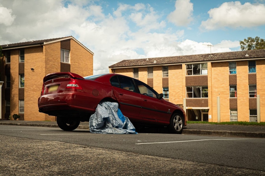 a car missing a wheel in front of a block of apartments