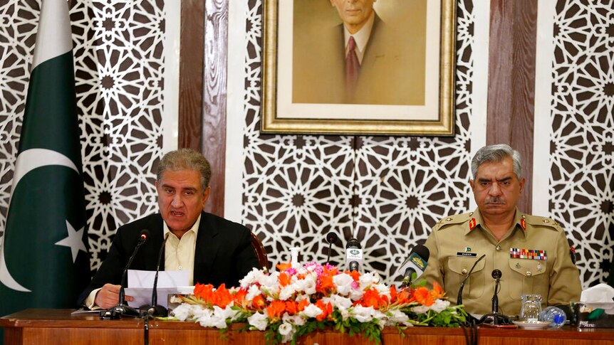 On a wooden desk, you view a man in a dark suit next to a man in light green military attire speaking into microphones.