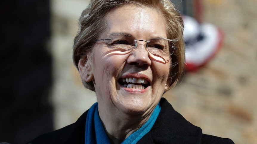 Elizabeth Warren, wearing a scarf and a coat, holds her hand to her chest and smiles.