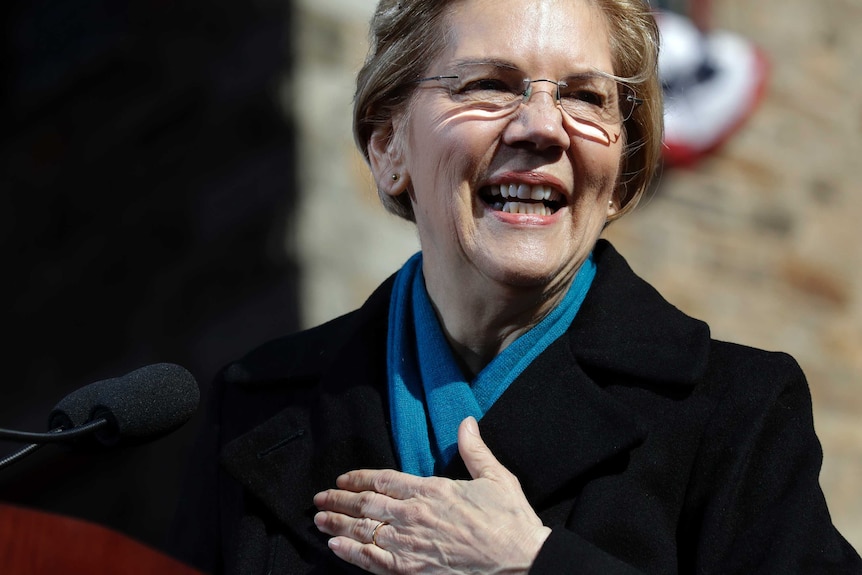 Elizabeth Warren, wearing a scarf and a coat, holds her hand to her chest and smiles.