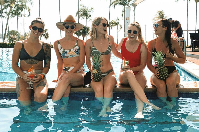 Five girls in bikinis laugh and hold cocktails as they sit poolside.