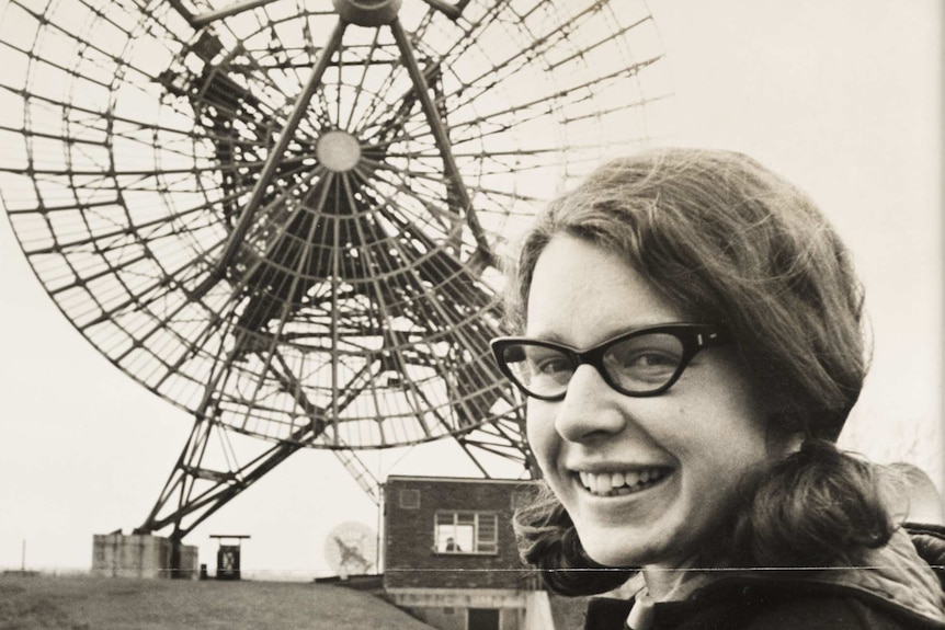 Jocelyn Bell with black horn-rimmed glasses, hair in pigtails, smiling, holding a scroll of paper. Radio receiver behind her