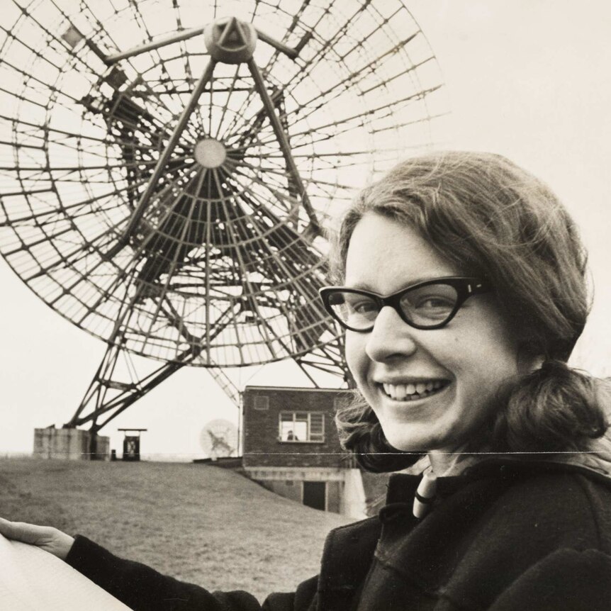 Jocelyn Bell with black horn-rimmed glasses, hair in pigtails, smiling, holding a scroll of paper. Radio receiver behind her