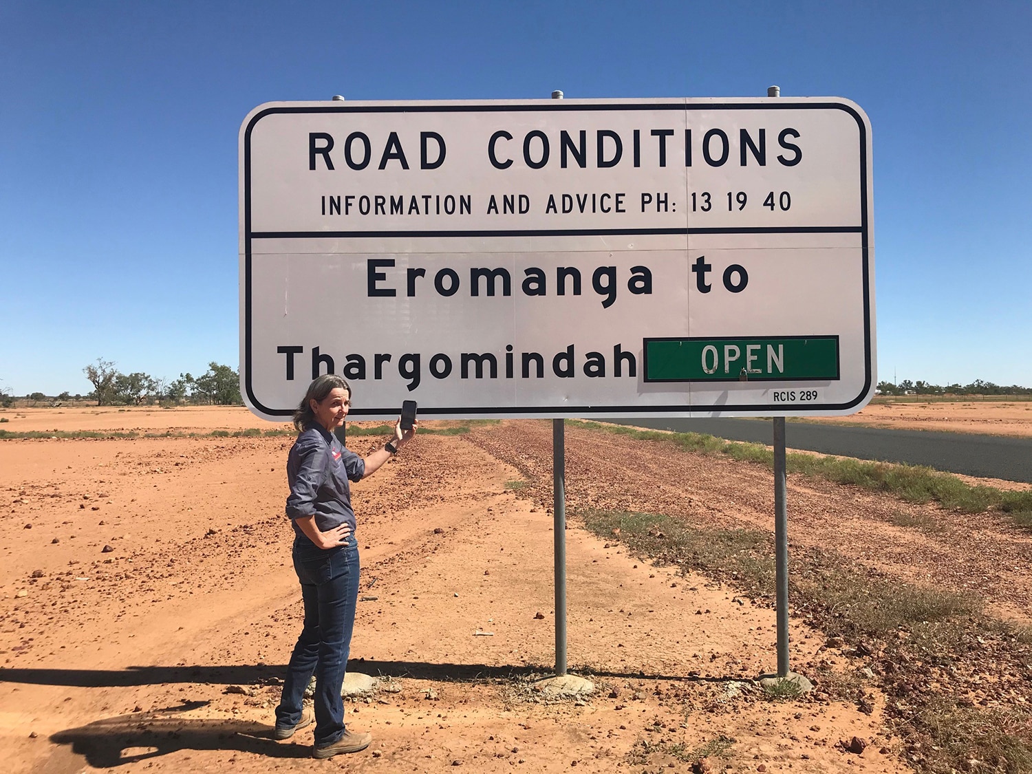 A woman in Eromanga looks at Google Maps on her phone, in front of a street sign 