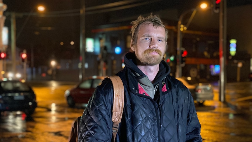 A man wearing a coat and carrying a backpack stands in front of cars and traffic lights at night.