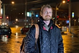 A man wearing a coat and carrying a backpack stands in front of cars and traffic lights at night.