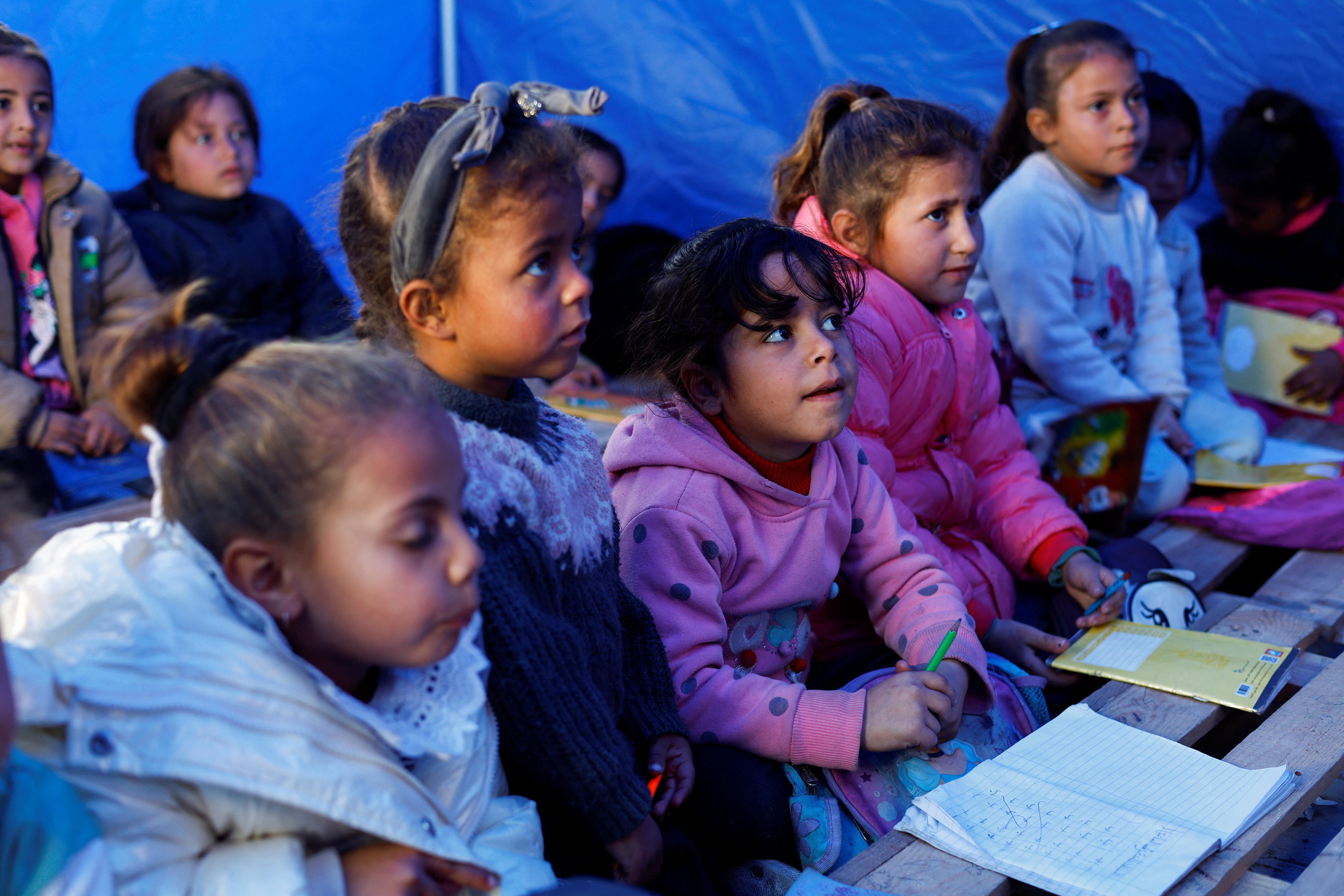 Young Palestinian girls sit on teh floor of a blue tent set up as a makeshift classroom. 