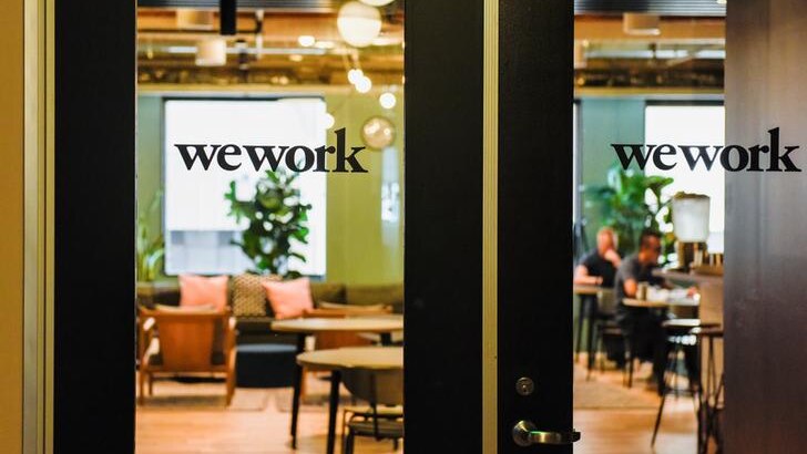 An office is seen behind two closed glass doors with text on them that read "wework". People are sat on chairs inside the room.