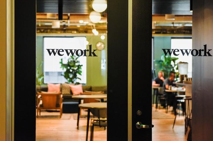 An office is seen behind two closed glass doors with text on them that read "wework". People are sat on chairs inside the room.