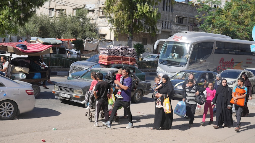 Palestinians walk down a dirt road carrying children and bags, cars are lined along the road