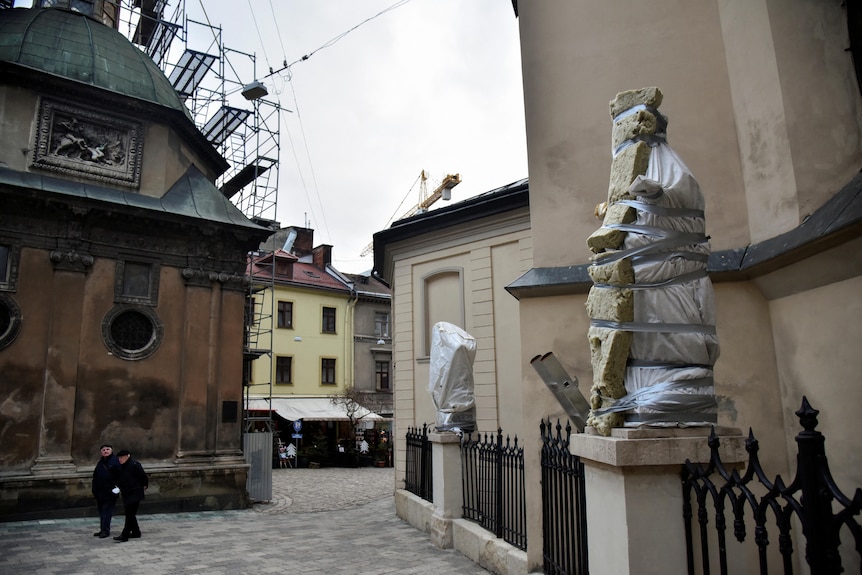 Two people walk past statues on the fence of a building which have been wrapped in foam, tape and fabric.