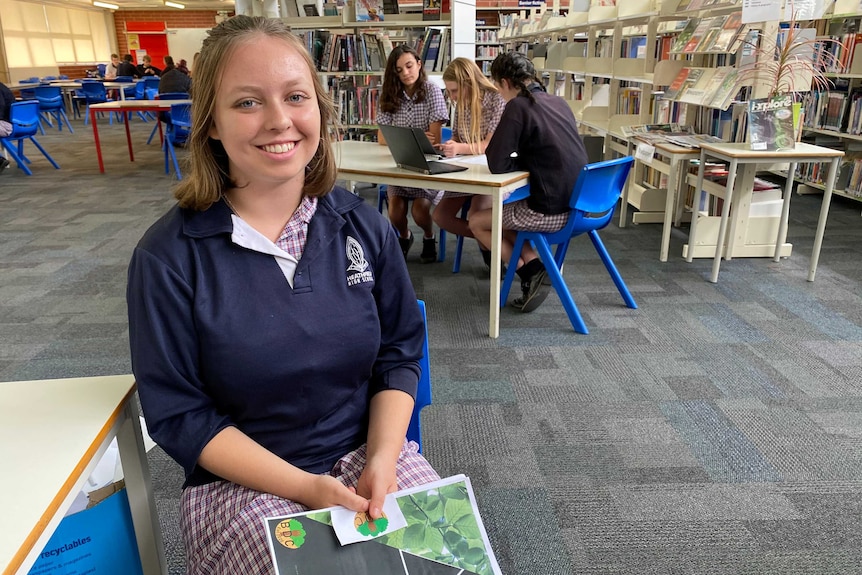 A female student smiling at the camera in the foreground with three female students working in the background in a library.