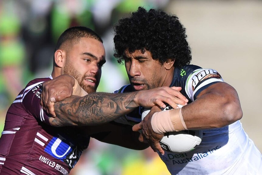 A male NRL player holds the ball with his left hand as he tries to fend off a tackle from an opposition player.
