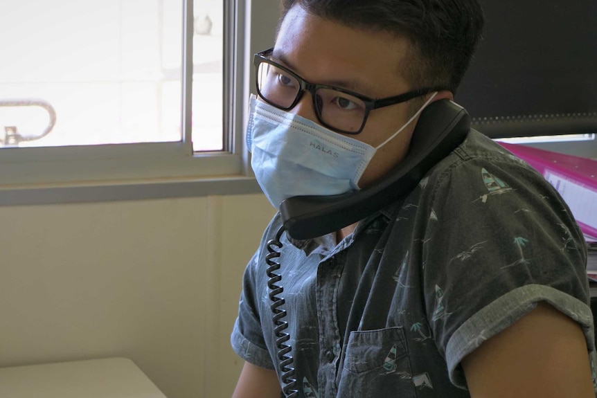 A man wearing a white face mask sits at a desk on the phone.