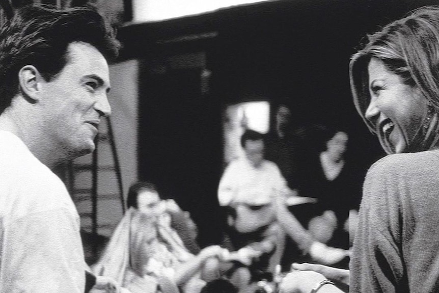 A black and white photo of Matthew Perry and Jennifer Aniston smiling at each other during a script reading session.