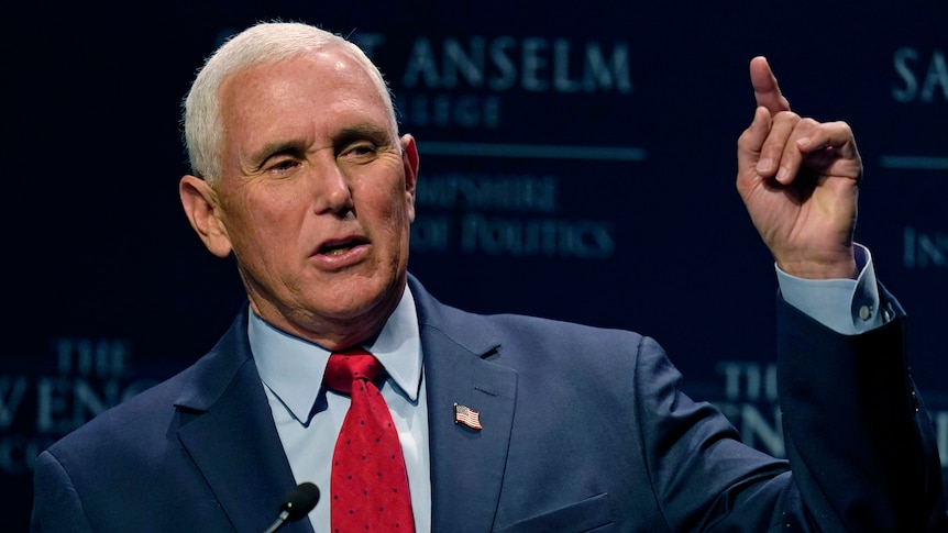 A white-haired man wearing a blue suit and red tie gestures while standing behind a microphone.