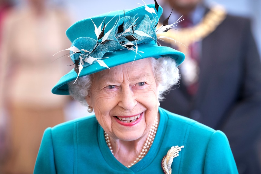 Queen Elizabeth smiles at the camera in a blue blazer and hat, July 1, 2021.