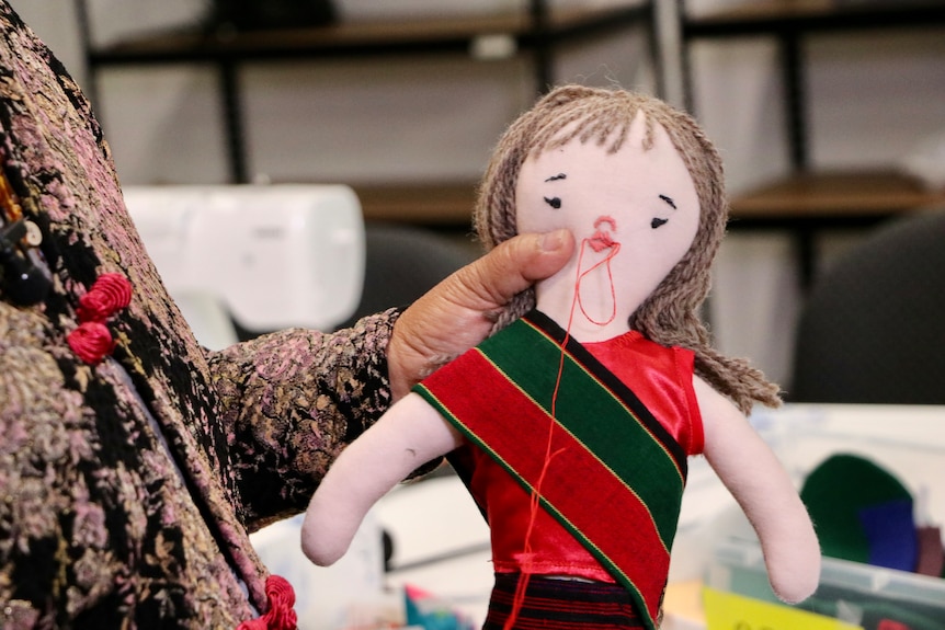 A close-up shot of a doll with its mouth being stitched by a woman's hands.