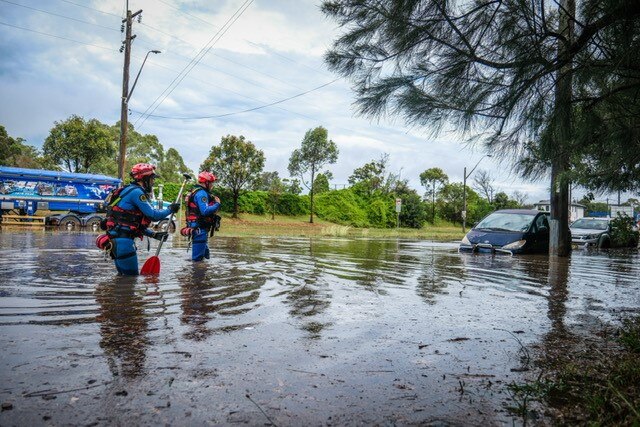 emergency service personnel in blue and red gear wade through knee deep water towards flooded cars 