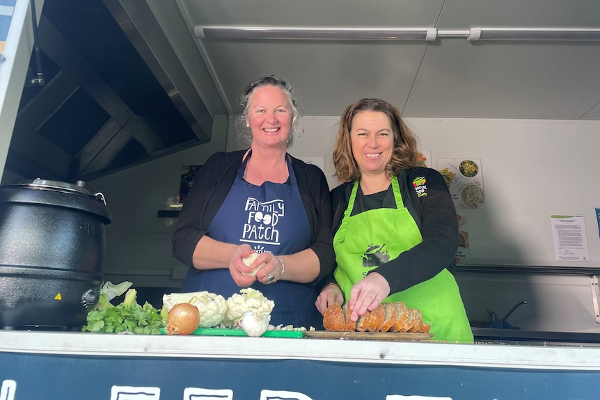 Two women cutting fresh fruit and veggies in a food truck smile down at the camera.