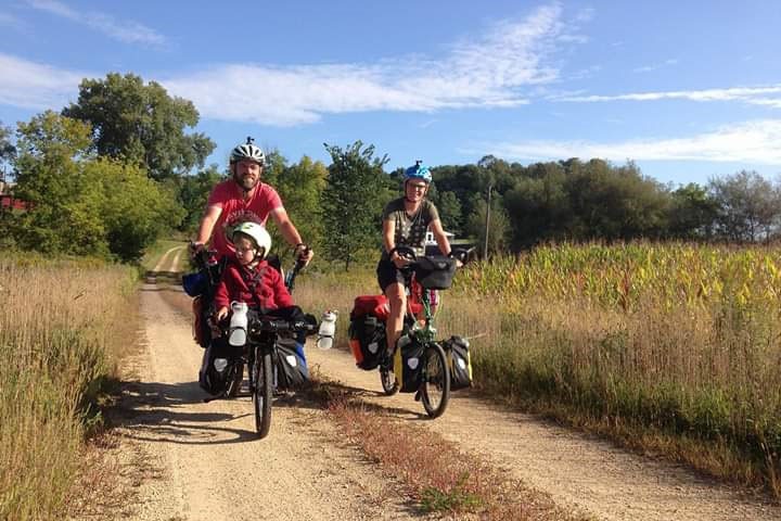 A man on a semi-recumbent bike with a young boy on the front ride on a dirt road with a woman