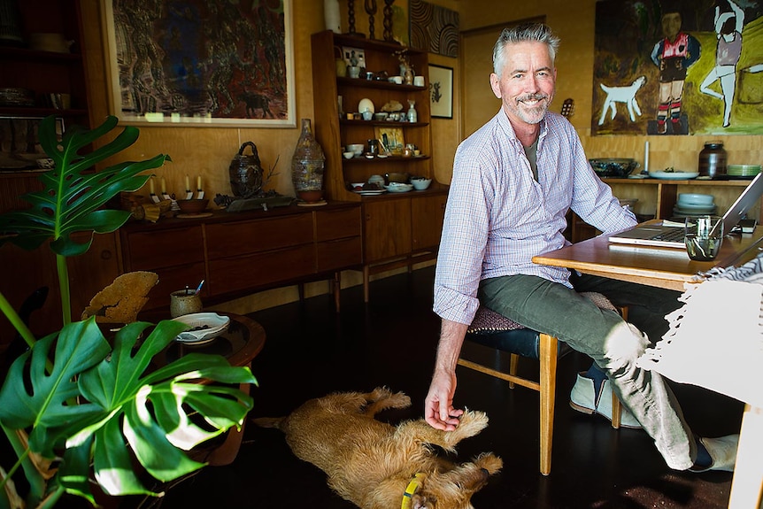 Man sits at computer, smiling and patting dog on the floor.