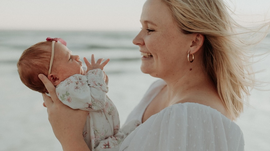 Close shot of lady smiling into the face of a baby she's holding in her hands with beach behind