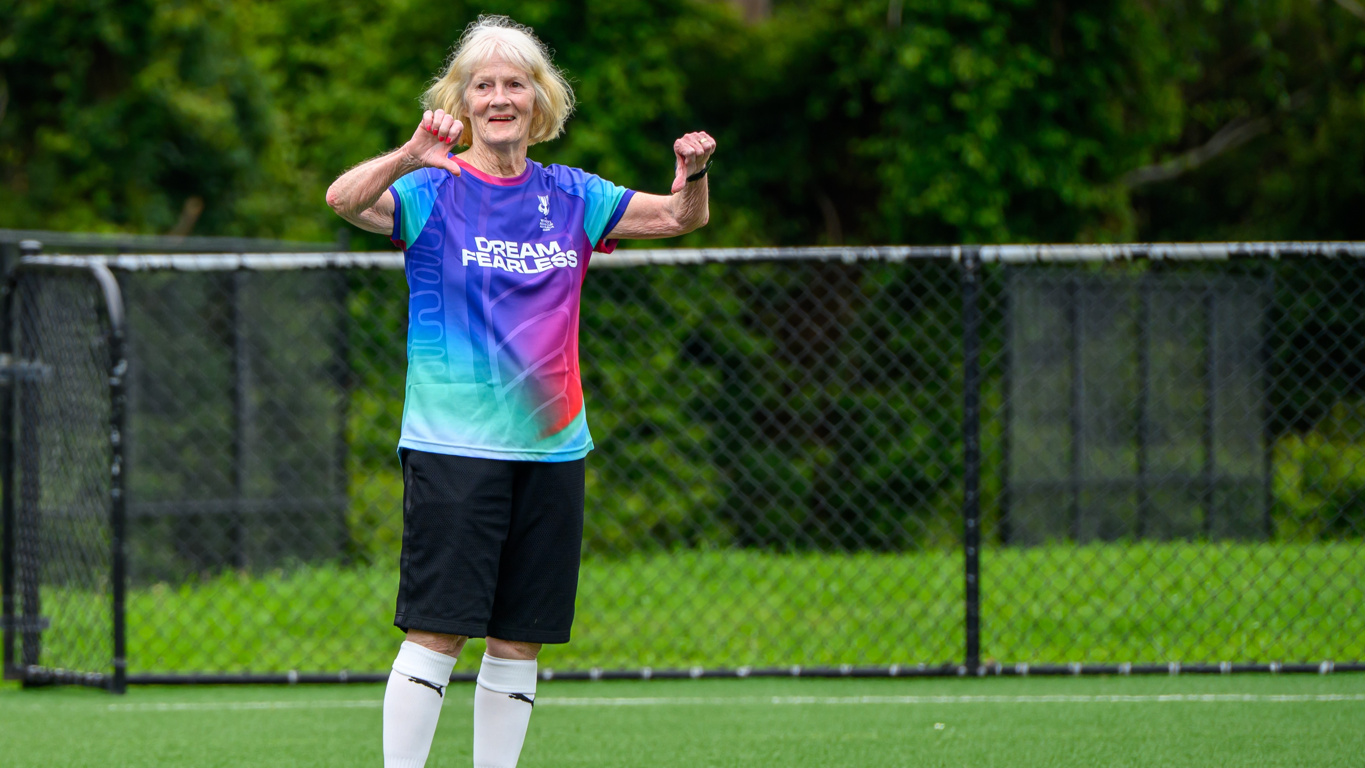 Uma mulher mais velha está jogando futebol, ela se levanta e sorri com os dois polegares para baixo.